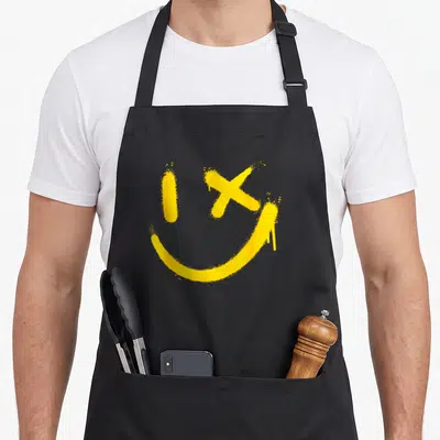 Tablier de cuisine noir avec un visage souriant jaune peint en aérographe, l'œil droit remplacé par une croix.