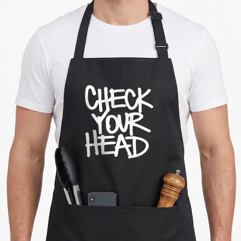 Tablier de cuisine noir avec l'inscription Check Your Head en lettres blanches stylisées type graffiti vintage.
