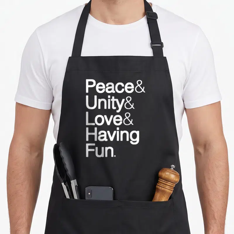 Tablier de cuisine noir avec le texte blanc Peace Unity Love Having Fun disposé verticalement sur le devant.