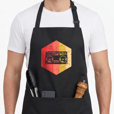 Tablier de cuisine noir avec une illustration hexagonale représentant une boombox vintage aux teintes orange et jaune, effet dégradé et usé.