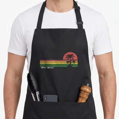 Tablier de cuisine noir avec un soleil rouge, bandes Rasta et palmiers gris stylisés sur fond uni.