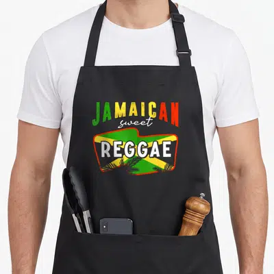 Tablier de cuisine noir avec l'inscription Jamaican Sweet Reggae sur fond de drapeau vert jaune et rouge orné de palmes.