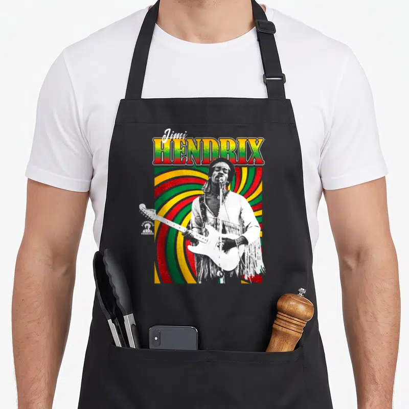 Tablier de cuisine noir avec une illustration colorée de Jimi Hendrix jouant de la guitare sur fond de motifs en spirale vert jaune et rouge.