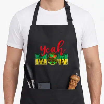 Tablier de cuisine noir avec inscription Yeah Mon Jamaica et illustration femme aux cheveux tressés portant des lunettes de soleil aux couleurs du drapeau jamaïcain.