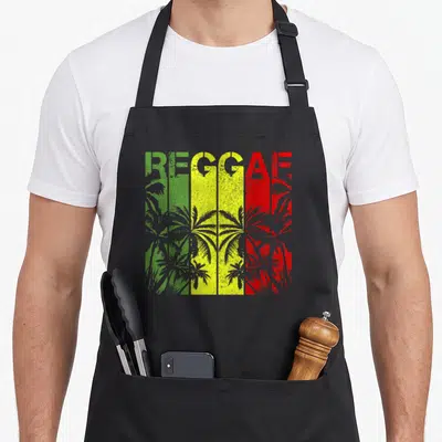 Tablier de cuisine noir avec linscription REGGAE aux couleurs rastafari et des palmiers gris en arrière-plan.