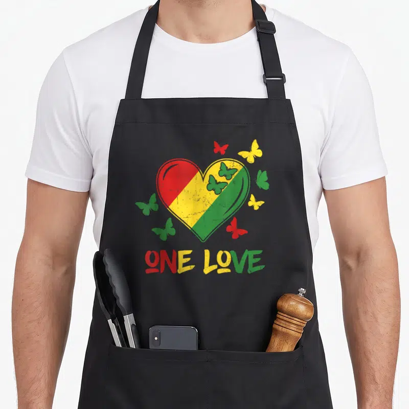 Tablier de cuisine noir avec un grand cœur tricolore Rasta et le texte One Love entouré de papillons colorés.