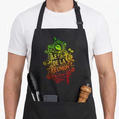 Tablier de cuisine noir avec une illustration colorée représentant l'Ile de la Réunion et des ornements végétaux aux teintes vert jaune et rouge.
