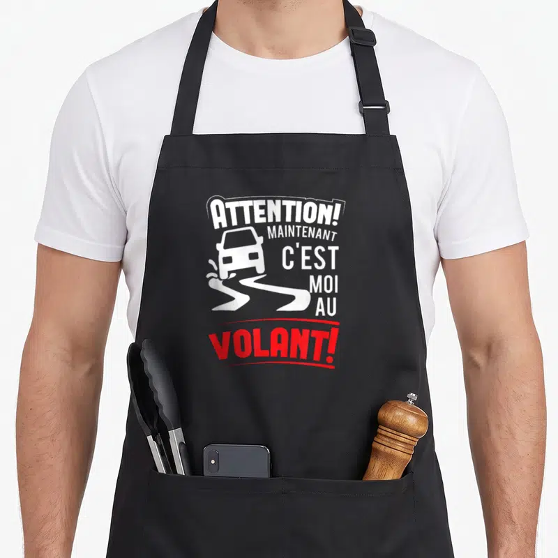 Tablier de cuisine noir avec une voiture blanche et le texte Attention maintenant c'est moi au volant en rouge et blanc.