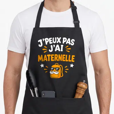 Tablier de cuisine noir avec inscription J'peux pas j'ai maternelle et illustration de sac à dos orange portant des lunettes.