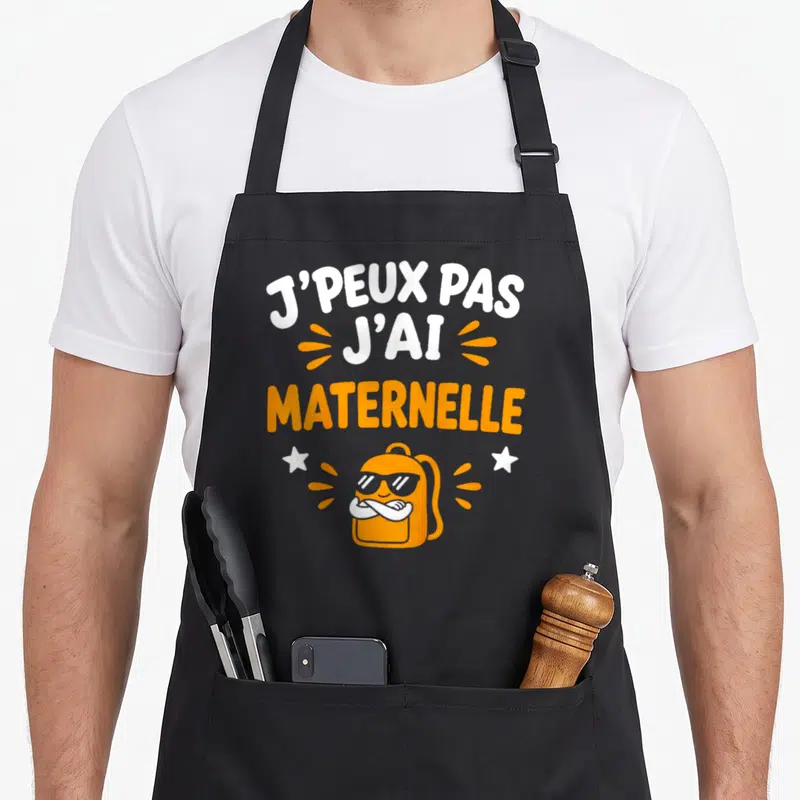 Tablier de cuisine noir avec inscription J'peux pas j'ai maternelle et illustration de sac à dos orange portant des lunettes.