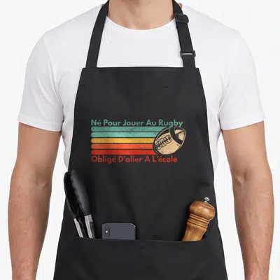 Tablier de cuisine noir avec inscription retro rugby et ballon ovale sur bandes colorées délavées.