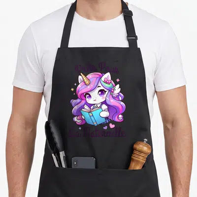 Tablier de cuisine noir avec une licorne rose et violette tenant un livre bleu, entourée d'étoiles et de cœurs.