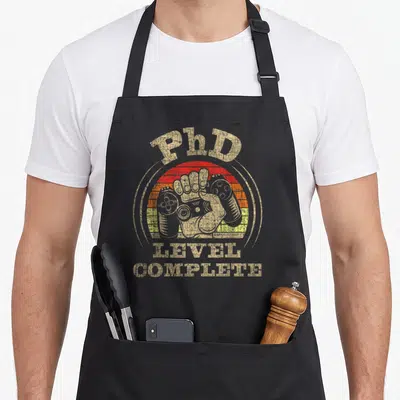 Tablier de cuisine noir avec illustration vintage PhD Level Complete et manette de jeu vidéo colorée.