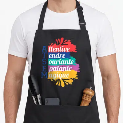 Tablier de cuisine noir avec le texte ATSEM et des traits de peinture colorés affichant des mots en anglais.