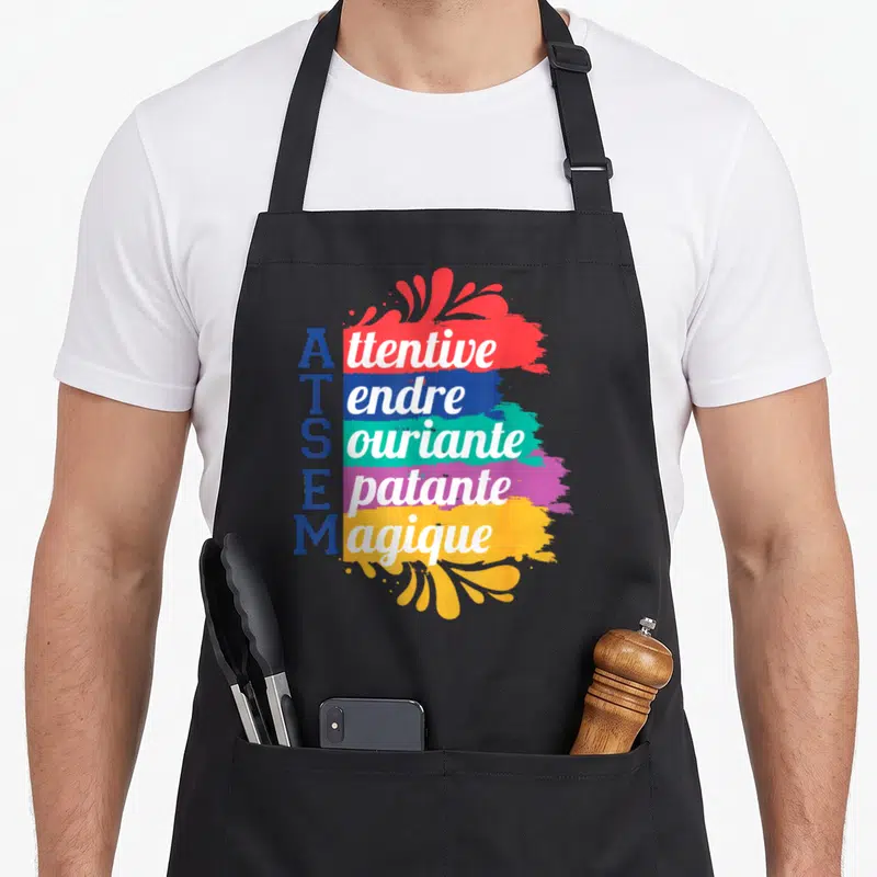 Tablier de cuisine noir avec le texte ATSEM et des traits de peinture colorés affichant des mots en anglais.