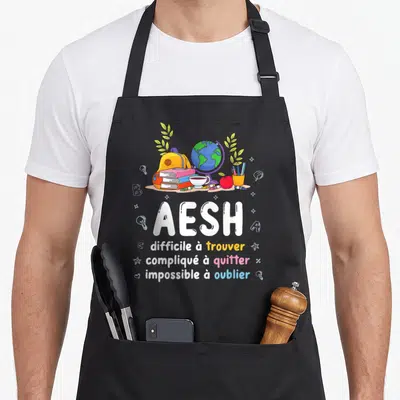 Tablier de cuisine noir avec le texte AESH difficile à trouver compliqué à quitter impossible à oublier entouré d'un sac à dos et d'un globe terrestre.