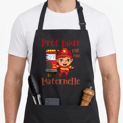 Tablier de cuisine noir avec un pompier cartoon courant devant une camionnette rouge et le texte Prêt pour la maternelle Pin Pon.