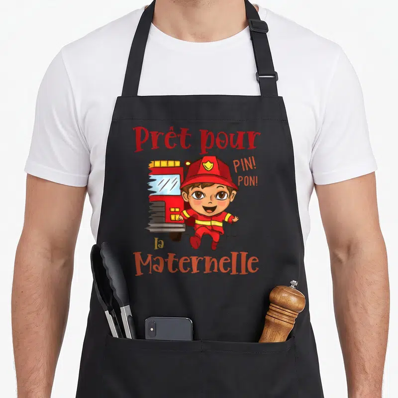 Tablier de cuisine noir avec un pompier cartoon courant devant une camionnette rouge et le texte Prêt pour la maternelle Pin Pon.