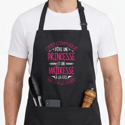 Tablier de cuisine noir avec l'inscription Cest compliqué dêtre une princesse et une maitresse en typographie rose et blanche.