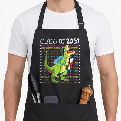 Tablier de cuisine noir avec un T-Rex vert portant sac à dos et casquette de graduation devant une liste de classes colorée.