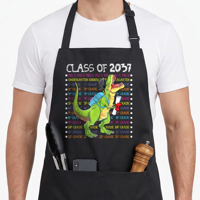 Tablier de cuisine noir avec un T-Rex vert portant sac à dos et casquette de graduation devant une liste de classes colorée.