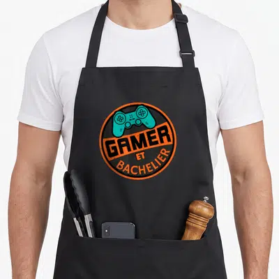 Tablier de cuisine noir avec logo circulaire orange affichant une manette turquoise et le texte GAMER ET BACHELIER en lettres noires.