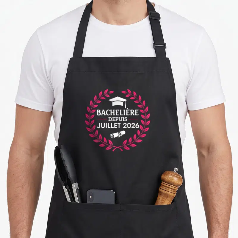 Tablier de cuisine noir avec inscription Bachelière Depuis Juillet 2026 entouré d'une couronne rose et d'un chapeau de graduation blanc.