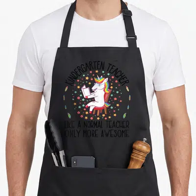 Tablier de cuisine noir avec une licorne lisant et le texte KINDERGARTEN TEACHER LIKE A NORMAL TEACHER ONLY MORE INCROYABLE entouré d'étoiles.