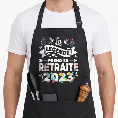 Tablier de cuisine noir avec inscription La Légende Prend Sa Retraite 2023 entourée de fleurs colorées et d'oiseaux blancs.