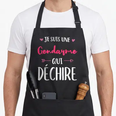 Tablier de cuisine noir avec inscription Je Suis Une Gendarme Qui Déchire en blanc et rose, orné de cœurs et d'une flèche.