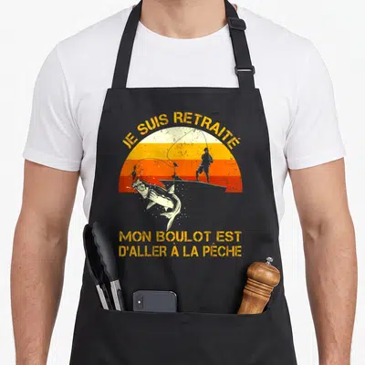 Tablier de cuisine noir avec illustration vintage d'un pêcheur et un poisson sautant devant un soleil orange, texte jaune en haut et en bas.