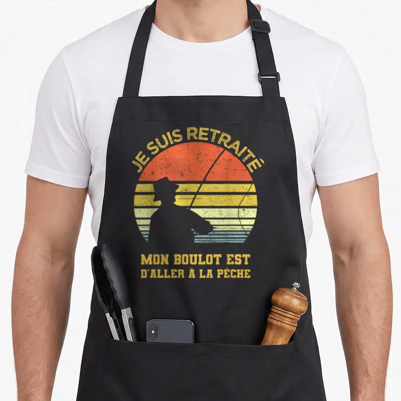 Tablier de cuisine noir avec silhouette pêcheur et texte Je suis retraité mon boulot est d'aller à la pêche sur fond de soleil coloré.