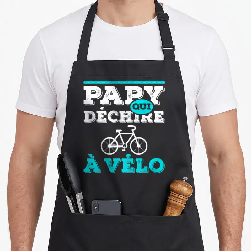 Tablier de cuisine noir avec le texte Papy qui déchire à vélo et un dessin de vélo blanc et turquoise.