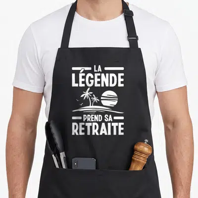 Tablier de cuisine noir avec inscription blanche La Légende Prend Sa Retraite sur fond de soleil et palmiers.