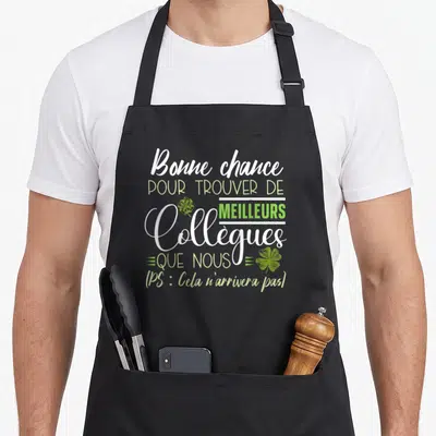 Tablier de cuisine noir avec l'inscription Bonne chance pour trouver de meilleurs collègues et des trèfles verts imprimés au centre.