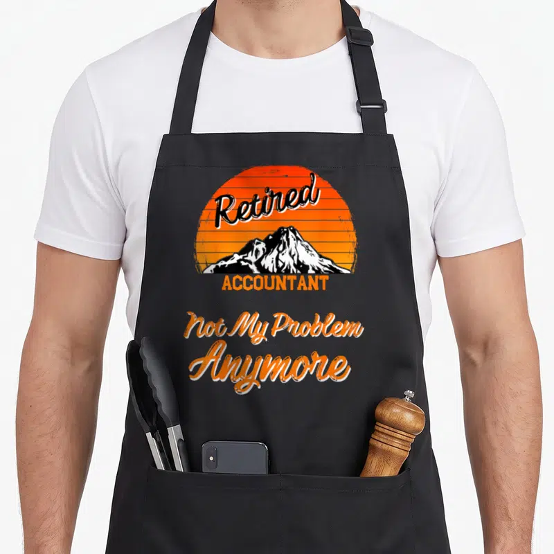 Tablier de cuisine noir avec motif soleil orange, montagne et texte Retired Accountant Not My Problem Anymore en lettres stylisées.