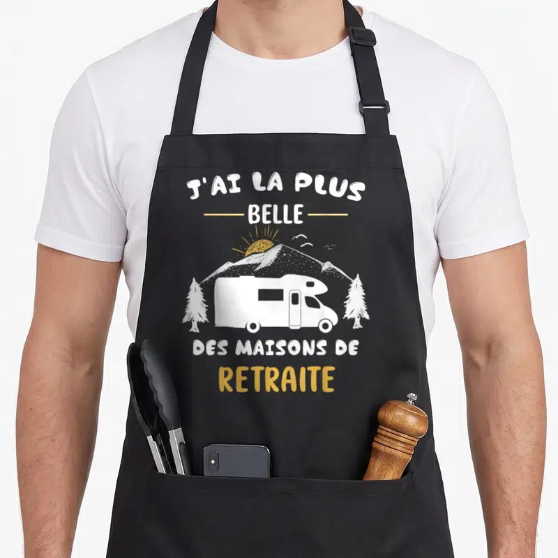 Tablier de cuisine noir avec illustration d'un camping-car blanc sous un soleil et des montagnes, texte en blanc et jaune.