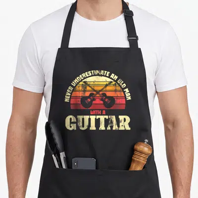 Tablier de cuisine noir avec deux guitares électriques croisées devant un soleil orange et rouge vintage.