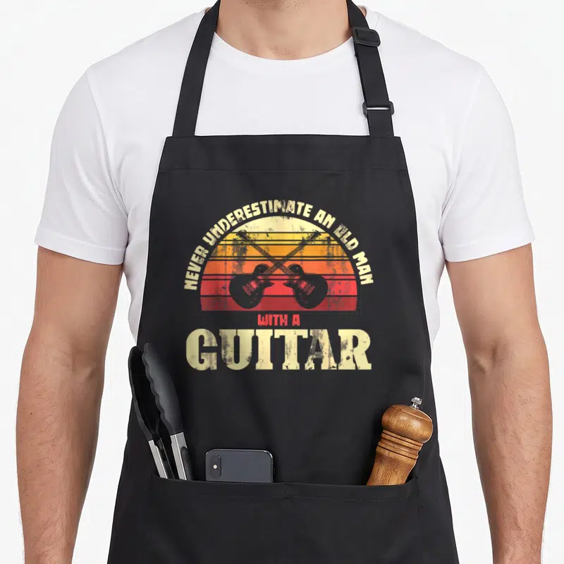 Tablier de cuisine noir avec deux guitares électriques croisées devant un soleil orange et rouge vintage.