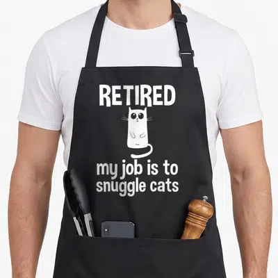 Tablier de cuisine noir avec illustration d'un chat blanc et texte retired my job is to snuggle cats en typographie blanche.