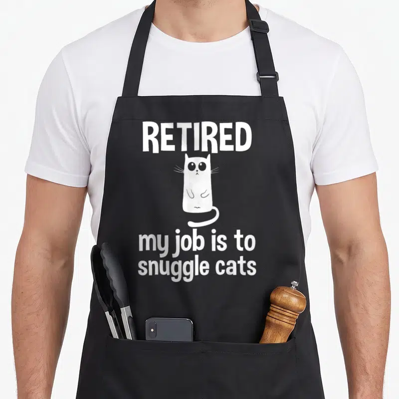 Tablier de cuisine noir avec illustration d'un chat blanc et texte retired my job is to snuggle cats en typographie blanche.