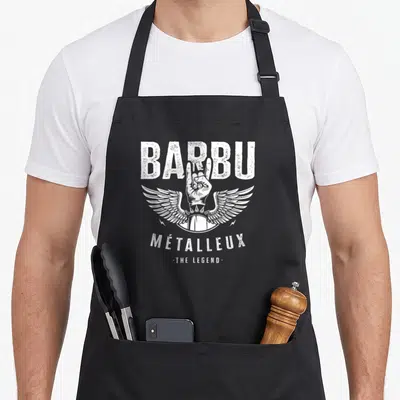 Tablier de cuisine noir avec poing de métal, ailes blanches et texte Barbu métalleux The Legend imprimé au centre.
