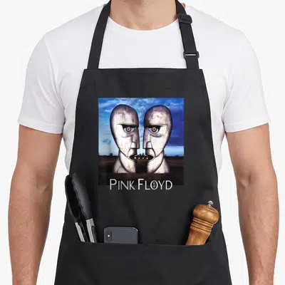 Tablier de cuisine noir avec deux têtes métalliques face à face et l'inscription Pink Floyd en blanc sur fond de ciel nuageux.