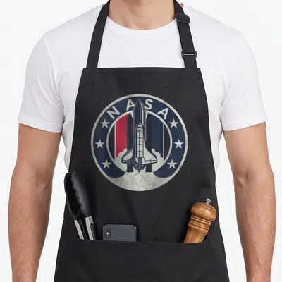 Tablier de cuisine noir avec logo circulaire NASA montrant une fusée blanche, des étoiles et un fond bleu vieilli.