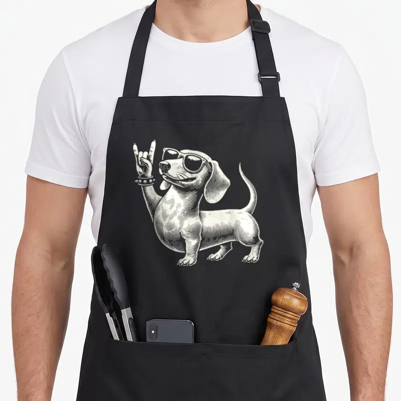 Tablier de cuisine noir avec un teckel dessiné en blanc portant des lunettes de soleil et levant la main en signe de rock.
