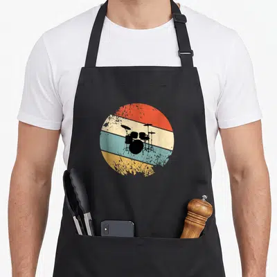 Tablier de cuisine noir avec illustration circulaire vintage représentant une batterie sur bandes colorées orange, bleu et jaune.