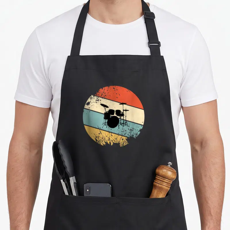 Tablier de cuisine noir avec illustration circulaire vintage représentant une batterie sur bandes colorées orange, bleu et jaune.