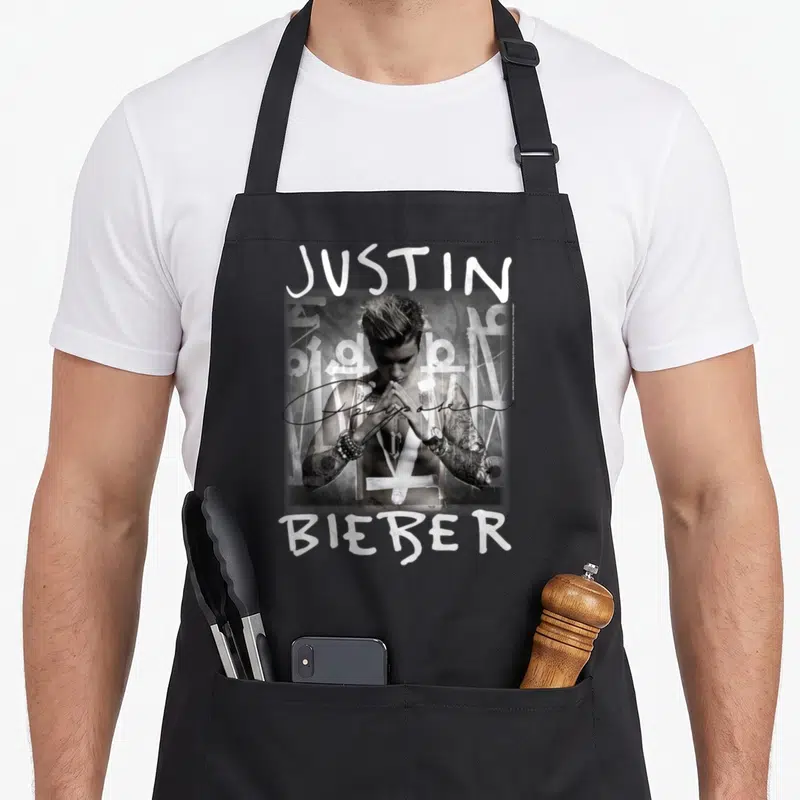 Tablier de cuisine noir avec portrait en noir et blanc de Justin Bieber, texte JUSTIN BIEBER et signature Purpose superposée.