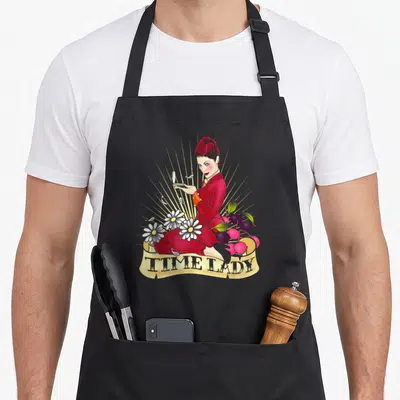 Tablier de cuisine noir avec illustration d'une femme en robe rouge tenant un miroir, des fleurs blanches et une bannière Time Lady.