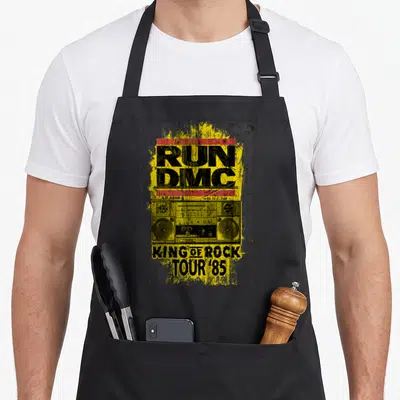 Tablier de cuisine noir avec illustration de boombox jaune et texte Run DMC King of Rock Tour 85 en style vintage.